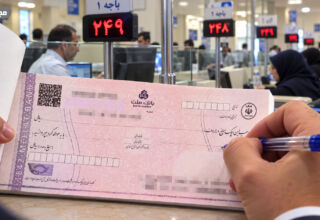 تعداد ضامن برای وام ودیعه مسکن در نمایی نزدیک از یک چک بانکی صورتی‌رنگ روی پیشخوان بانک، در حالی که فردی با خودکار آبی در حال تکمیل آن است و فضای داخلی شعبه بانک با باجه‌ها، کارکنان و تابلوهای نوبت‌دهی در پس‌زمینه دیده می‌شود.