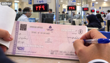 تعداد ضامن برای وام ودیعه مسکن در نمایی نزدیک از یک چک بانکی صورتی‌رنگ روی پیشخوان بانک، در حالی که فردی با خودکار آبی در حال تکمیل آن است و فضای داخلی شعبه بانک با باجه‌ها، کارکنان و تابلوهای نوبت‌دهی در پس‌زمینه دیده می‌شود.